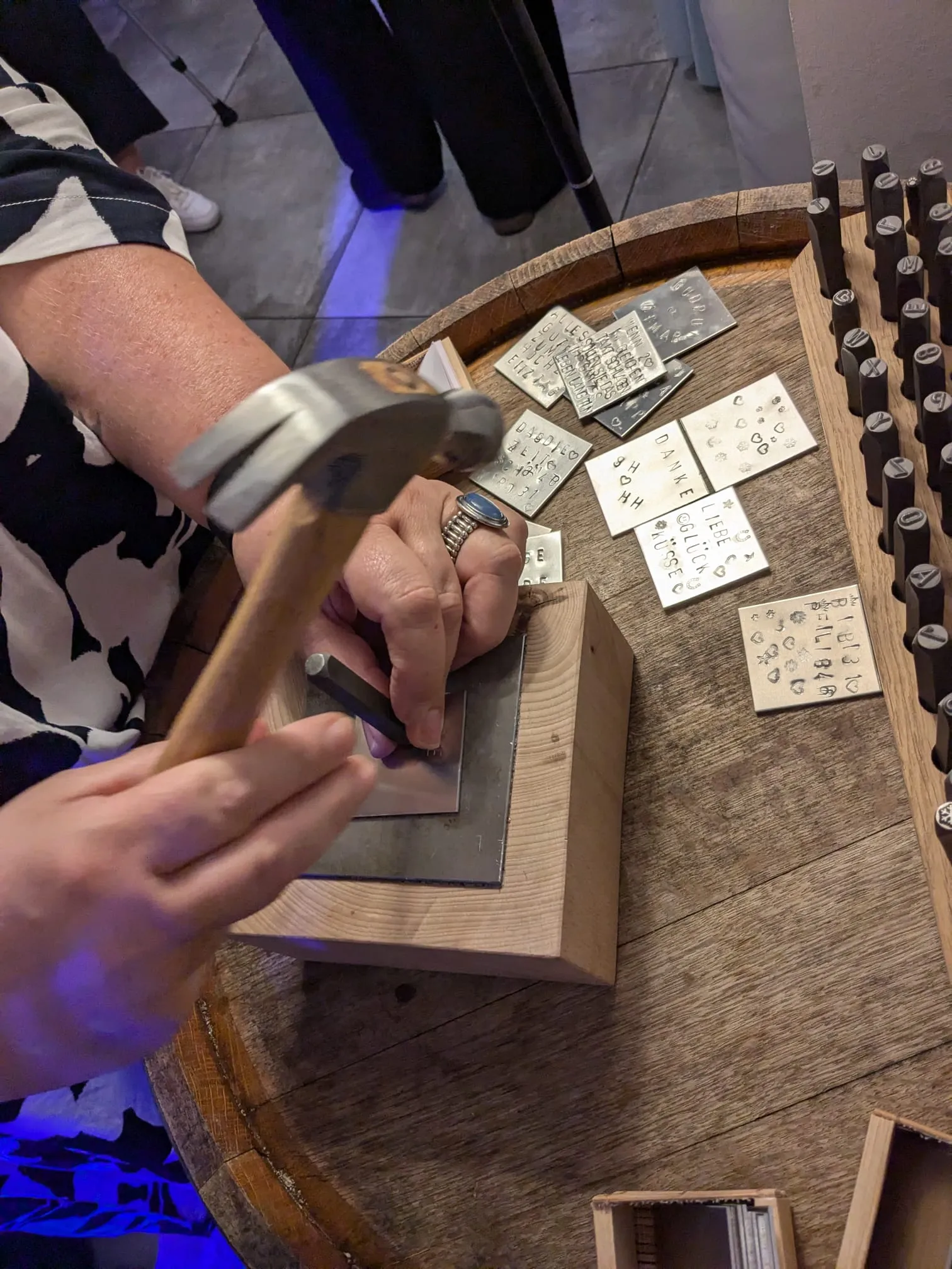 Hochzeitsgast hämmert ein Wort in ein Metallplättchen, umgeben von fertig geprägten Gäste-Plättchen beim Wortschmiede-Ritual