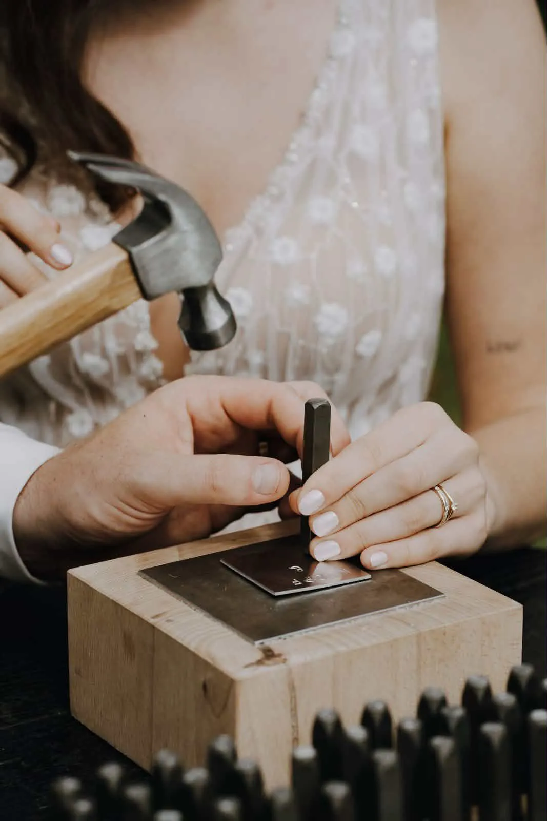 Braut im Blumenkleid und Bräutigam hämmern gemeinsam ein Wort in die Metallplatte beim Wortschmiede-Ritual