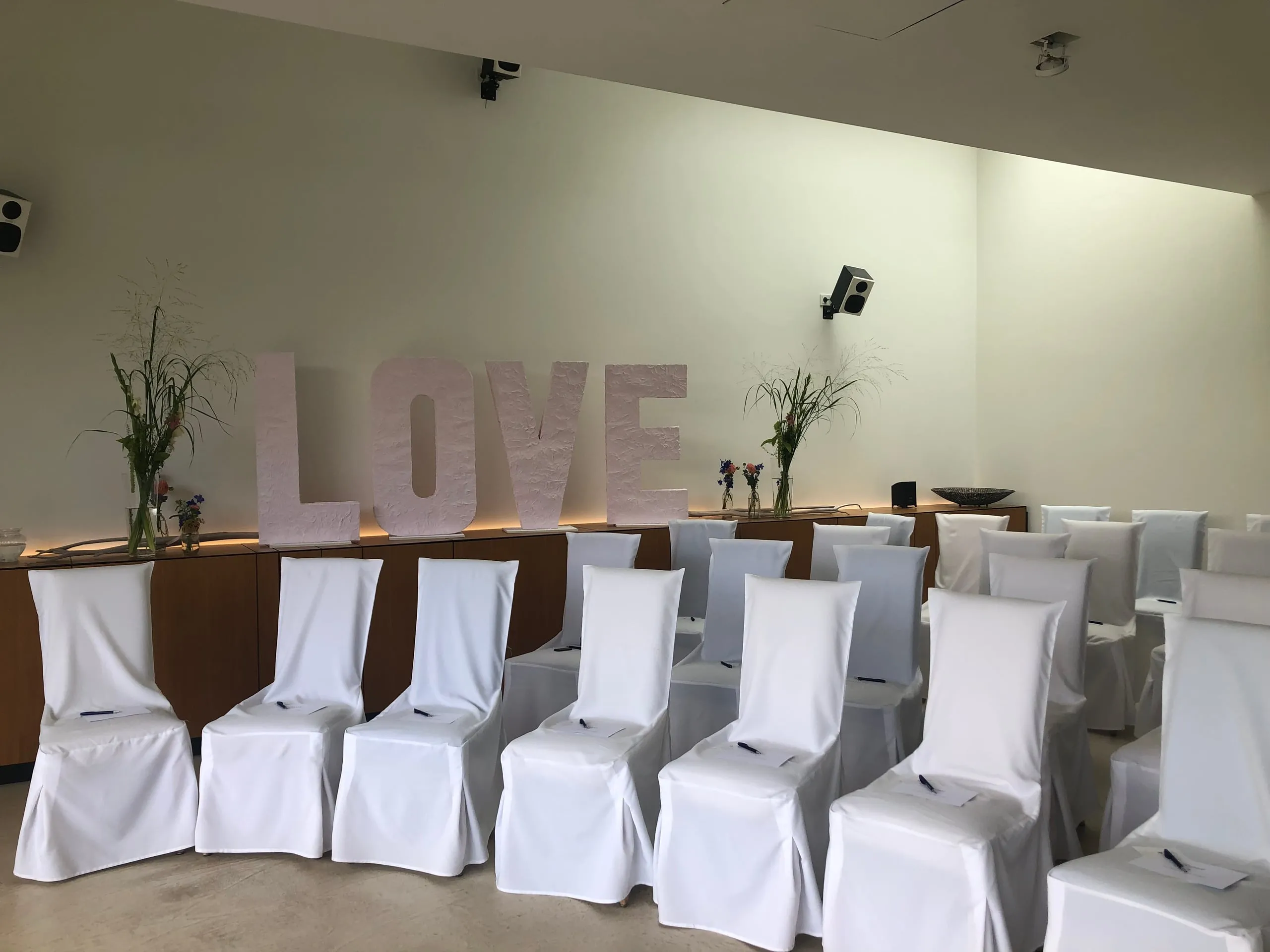 LOVE-Buchstaben im Zeremoniesaal mit weißen Stuhlhussen und Wildblumen in Glasvasen