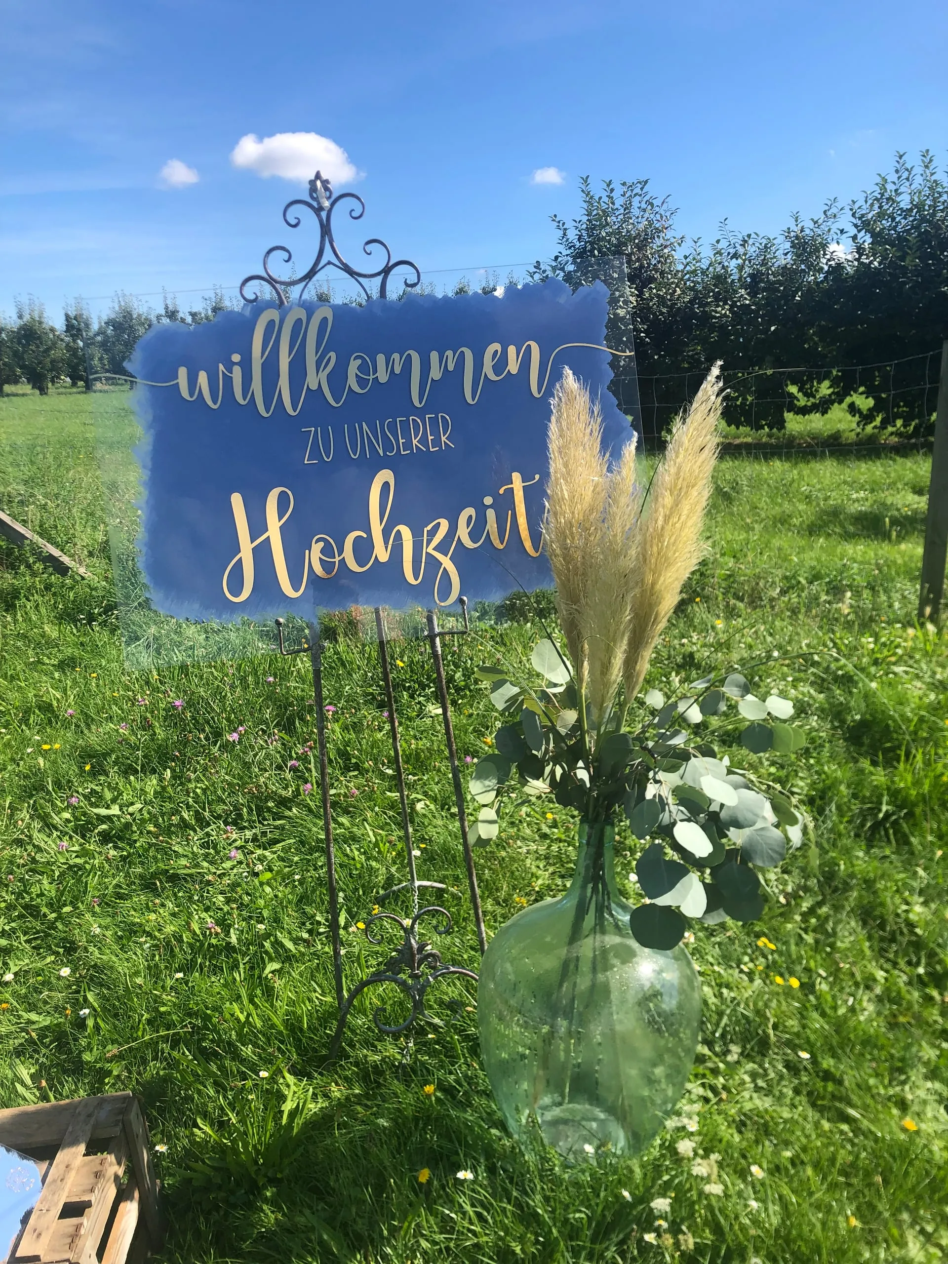 Blaues Willkommensschild mit goldener Schrift und Pampasgras bei einer Hochzeit