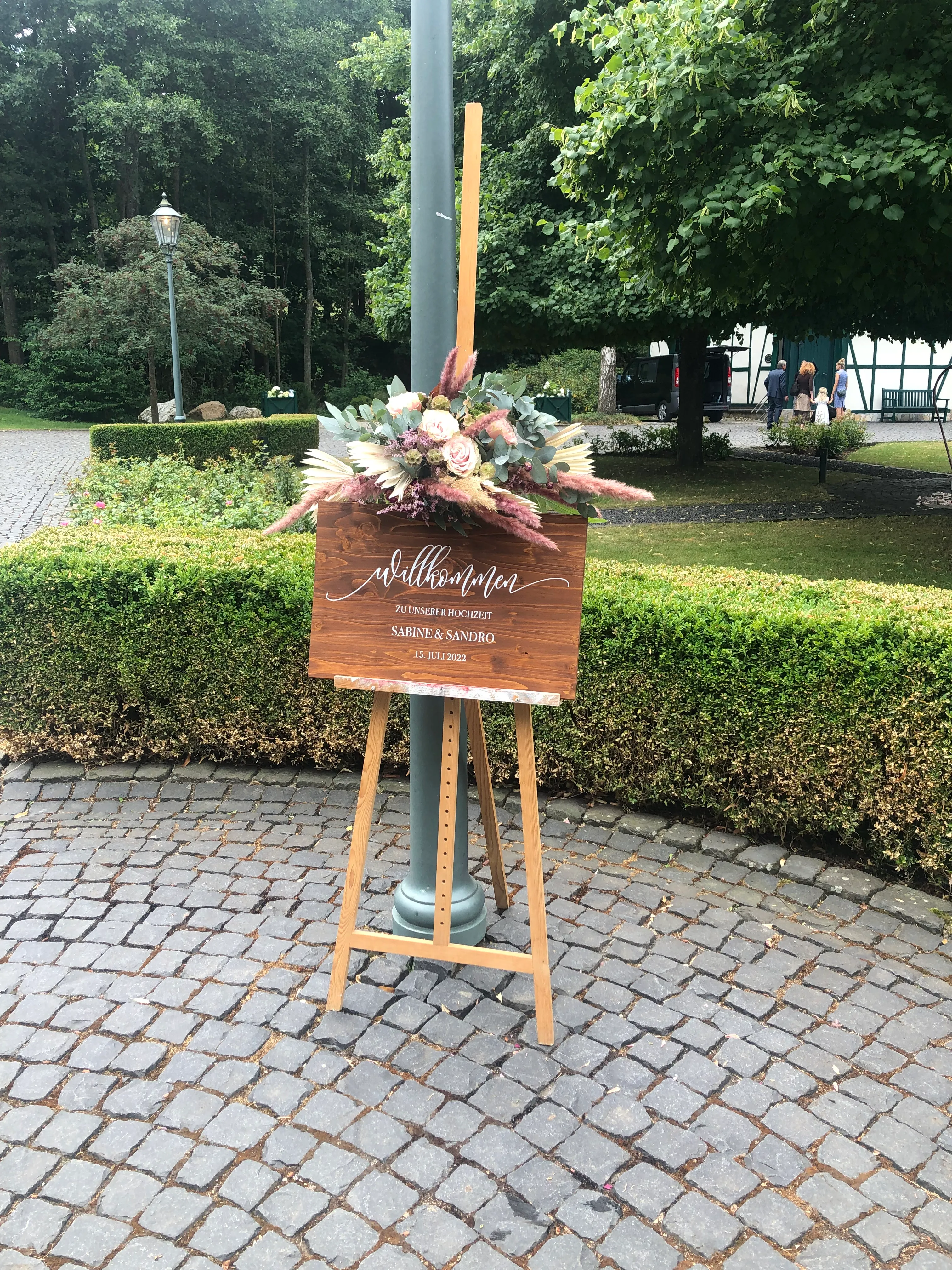 Willkommensschild aus Holz mit Blumenarrangement auf einer Staffelei bei einer freien Trauung